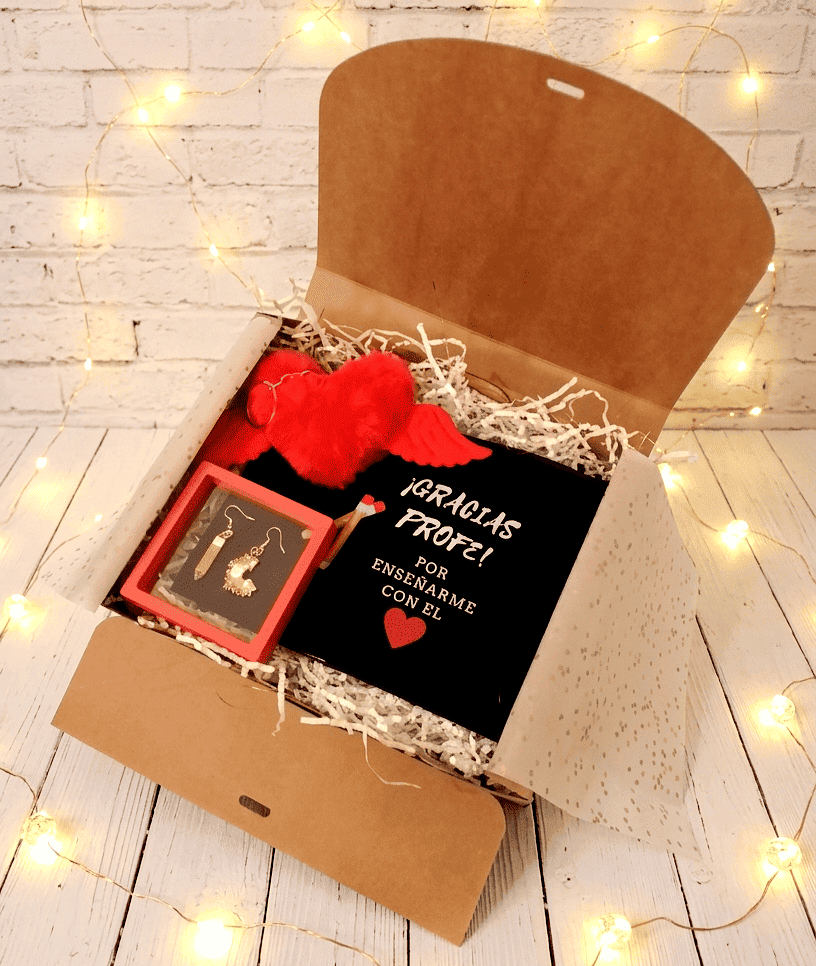Caja de regalo abierta vista desde arriba mostrando un neceser negro con frase de agradecimiento para profesor en español, con letra blanca y dibujo de corazón rojo y dos lapiceros. En la parte superior izquierda puede verse un llavero en forma de corazón rojo de peluche con alas, justo debajo pueden verse unos originales pendientes, uno es un lapicero y el otro una viruta de sacapuntas en caja de exhibición con marco rojo, todo sobre una superficie de madera blanca con luces led. 