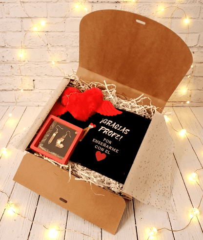 Caja de regalo abierta vista desde arriba mostrando un neceser negro con frase de agradecimiento para profesor en español, con letra blanca y dibujo de corazón rojo y dos lapiceros. En la parte superior izquierda puede verse un llavero en forma de corazón rojo de peluche con alas, justo debajo pueden verse unos originales pendientes, uno es un lapicero y el otro una viruta de sacapuntas en caja de exhibición con marco rojo, todo sobre una superficie de madera blanca con luces led. 