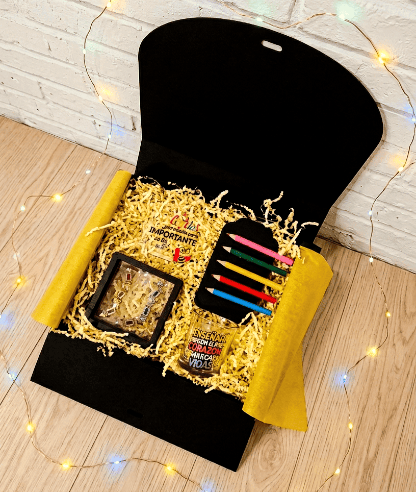 Caja de regalo vista desde arriba que contiene empezando por la parte superior izquierda una placa acrilica con frase de agradecimiento en español y con detalles coloridos, a su lado hay cinco pinzas en forma de pintutas de madera en diferentes colores presentadas en cartulina negra, debajo puede verse una vela en portavelas de cristal con diseño multicolor de frase inspiradora en español, a la izquierda de la vela pueden verse unos pendientes con colgantes de pequeños crayones de madera multicolor.