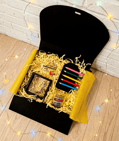 Caja de regalo vista desde arriba que contiene empezando por la parte superior izquierda una placa acrilica con frase de agradecimiento en español y con detalles coloridos, a su lado hay cinco pinzas en forma de pintutas de madera en diferentes colores presentadas en cartulina negra, debajo puede verse una vela en portavelas de cristal con diseño multicolor de frase inspiradora en español, a la izquierda de la vela pueden verse unos pendientes con colgantes de pequeños crayones de madera multicolor.