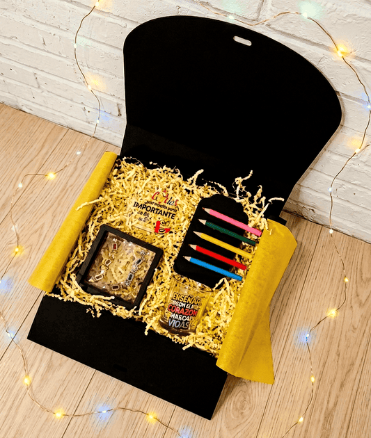 Caja de regalo vista desde arriba que contiene empezando por la parte superior izquierda una placa acrilica con frase de agradecimiento en español y con detalles coloridos, a su lado hay cinco pinzas en forma de pintutas de madera en diferentes colores presentadas en cartulina negra, debajo puede verse una vela en portavelas de cristal con diseño multicolor de frase inspiradora en español, a la izquierda de la vela pueden verse unos pendientes con colgantes de pequeños crayones de madera multicolor.