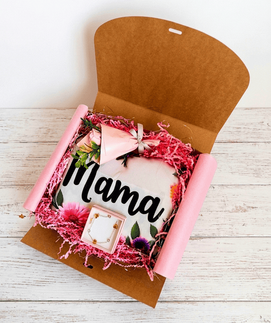 Caja de regalo abierta que contiene una Bolsa Tote plegada con la palabra Mama sobre fondo blanco y flores alrededor, en la parte superior de la caja hay una rosa eterna con envoltorio de papel rosa, en la parte inferior de la caja se puede ver una cajita con una pulsera de Acero dorado. Todo descansa sobre virutas de papel rosa, sobre una superficie de madera blanca.