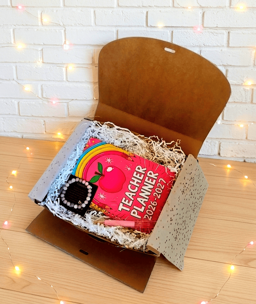 Caja de regalo abierta vista desde arriba en la que se puede ver una agenda colocada en horizontal de color fucsia con un diseño pop-retro colorido. En la parte inferior izquierda hay una pulsera de cuentas en tonos rosados presentada en tarjeta negra con dedicatoria de letras doradas a su lado se encuentra un boligrafo rosa y acrilico con flores secas en su interior. La caja está sobre una superficie de madera clara y luces Led.