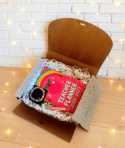 Caja de regalo abierta vista desde arriba en la que se puede ver una agenda colocada en horizontal de color fucsia con un diseño pop-retro colorido. En la parte inferior izquierda hay una pulsera de cuentas en tonos rosados presentada en tarjeta negra con dedicatoria de letras doradas a su lado se encuentra un boligrafo rosa y acrilico con flores secas en su interior. La caja está sobre una superficie de madera clara y luces Led.