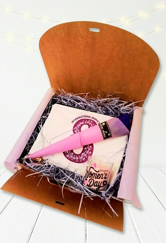 Caja de regalo abierta con virutas de papel malva mostrando un neceser con un 8 en morado, una rosa morada con envoltorio en color malva y una vela rosa con la frase "Happy women's day" sobre superficie de madera blanca.