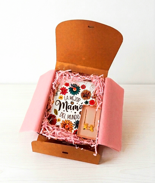 Caja de regalo abierta con un necesrde estampado floral con la frase " La mejor madre del mundo" junto a una caja con un colgante dorado de dos manos formando un corazón  sobre una superficie de madera blanca. 