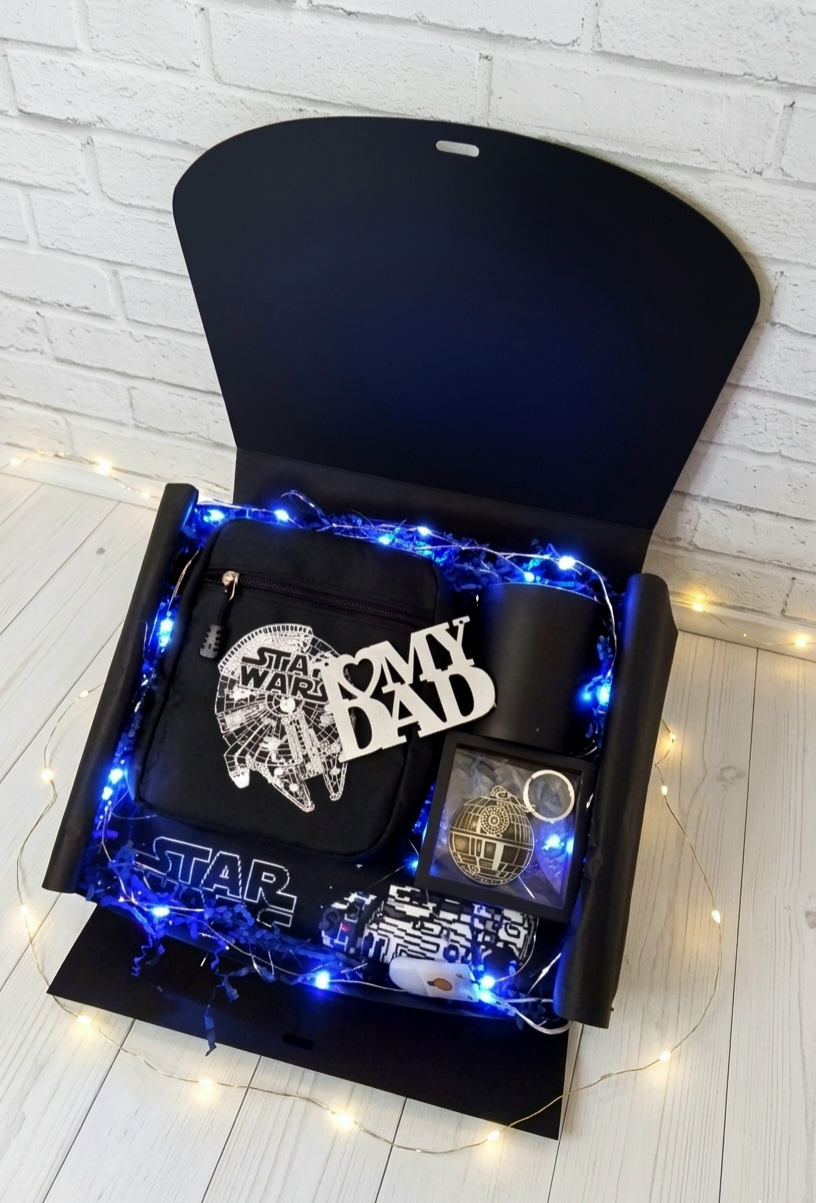 Caja de regalo negra abierta para el dia del padre con bolso bandolera del Halcon Milenario, llavero, taza negra, protector de cinturón sobre virutas de papel azul y luces led 