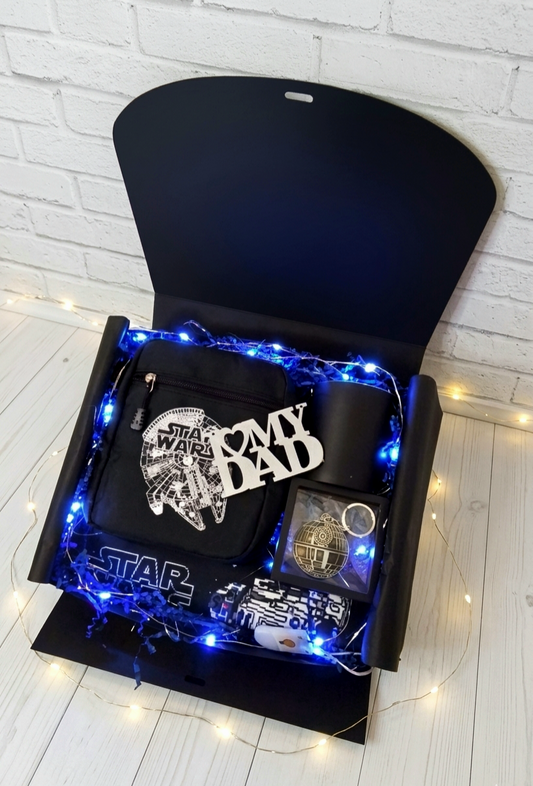 Caja de regalo negra abierta para el dia del padre con bolso bandolera del Halcon Milenario, llavero, taza negra, protector de cinturón sobre virutas de papel azul y luces led 