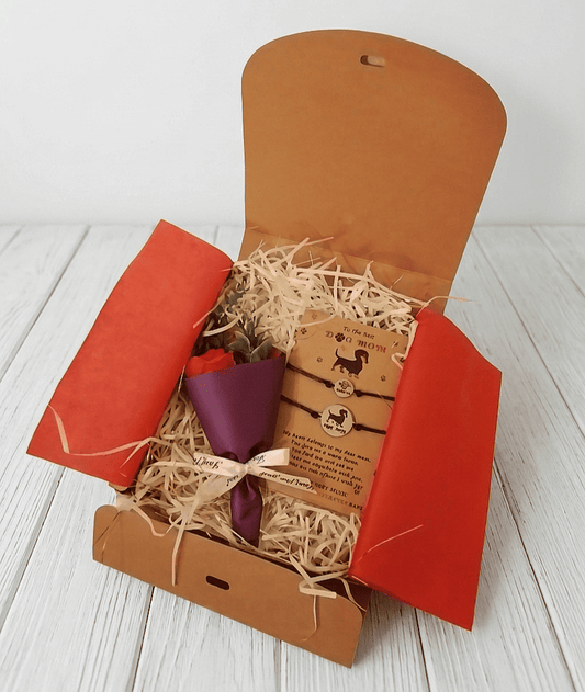 Vista superior de una caja de regalo abierta sobre madera blanca, contiene una Rosa Eterna de jabón de color rojo con envoltorio de papel satinado e color burdeos junto a una tarjeta de papel kraft que contiene un set de pulseras : una con un perro grabado y la otra con una huella de pata.