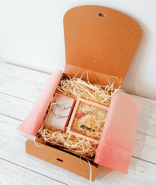 Vista superior de una caja de regalo abierta con virutas de papel blanco que contiene un set de pulseras de corazón y un llavero circular con la palabra Mamá sobre superficie de madera blanca. 