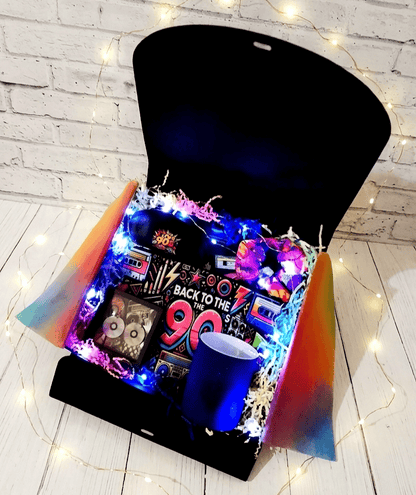 Caja de regalo abierta vista desde arriba, contiene articulos y objetos con diseño de los 90, con mucho colorido, logotipos y textos de los 90 en colores fluor y con luces led en su interior, sobre una superficie de madera blanca y pared blanca de ladrillo visto.