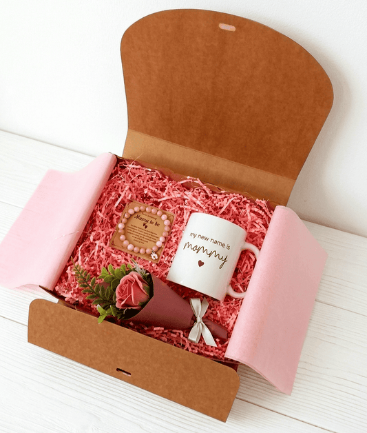 Caja de regalo abierta vista desde arriba mostrando una taza blanca de ceramica con mensaje en inglés de dia de la madre, una pulsera de cuentas en colores pastel y una rosa eterna sobre virutas de papel rosa.