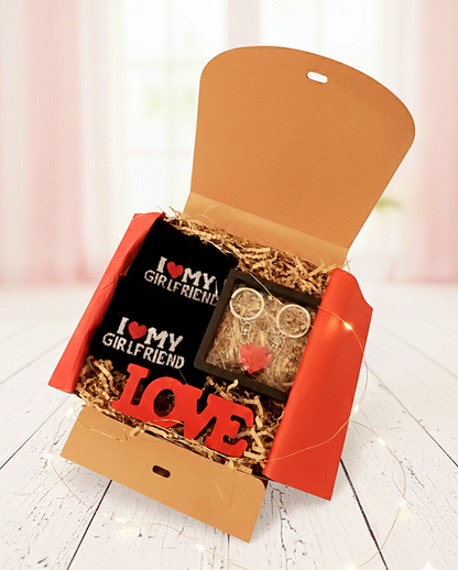 Caja de regalo abierta mostrando un llavero de piezas lego en color rojo formando un corazón, un par de calcetines negros con la frase "I ❤️ My Girlfriend" y un letrero de madera en color rojo con la palabra LOVE, sobre superficie blanca con luces led. 