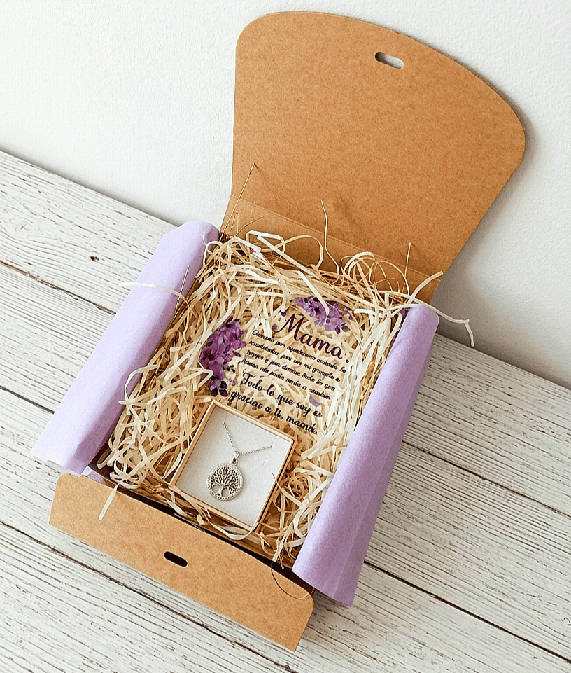 Caja de regalo Kraft abierta vista desde arriba, contiene una placa de metacrilato en forma de corazón con texto emotivo de dia de la madre, decorado con flores lilas y un colgate de Acero en color plateado de arbol de la vida sobre una  superficie de madera blanca. 