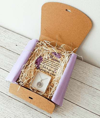 Caja de regalo Kraft abierta vista desde arriba, contiene una placa de metacrilato en forma de corazón con texto emotivo de dia de la madre, decorado con flores lilas y un colgate de Acero en color plateado de arbol de la vida sobre una  superficie de madera blanca. 