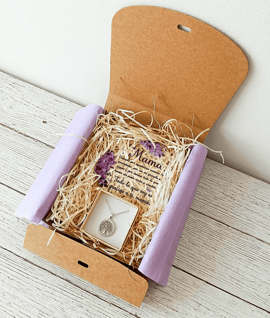 Caja de regalo Kraft abierta vista desde arriba, contiene una placa de metacrilato en forma de corazón con texto emotivo de dia de la madre, decorado con flores lilas y un colgate de Acero en color plateado de arbol de la vida sobre una  superficie de madera blanca. 