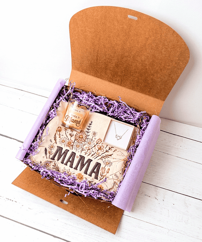 Caja de regalo abierta vista desde arriba, sobre una superficie de madera blanca,  se puede ver en su interior un neceser de diseño floral con la palabra Mamá, en la parte superior izquierda hay una vela decorativa con un emotivo mensaje de dia de la madre, a su derecha se puede ver una cajita de joyería rosa con el fondo blanco y en su interior hay un colgante plateado de Acero compueto por un simbolo infinito entrelazado con un corazón .
