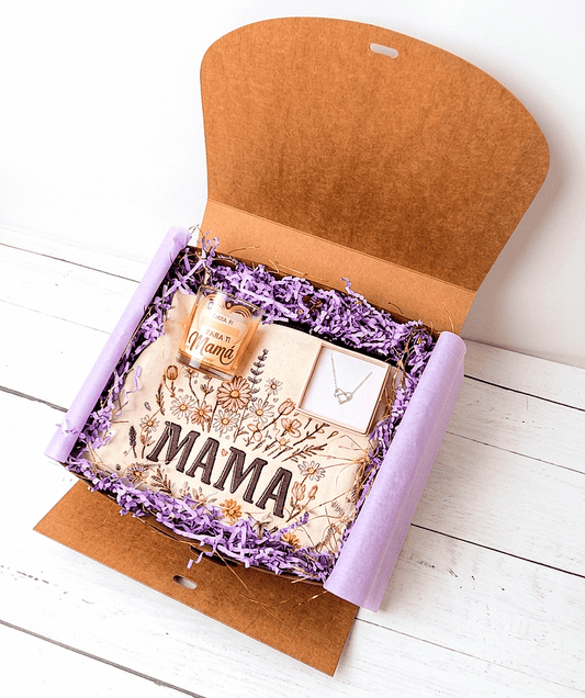 Caja de regalo abierta vista desde arriba, sobre una superficie de madera blanca,  se puede ver en su interior un neceser de diseño floral con la palabra Mamá, en la parte superior izquierda hay una vela decorativa con un emotivo mensaje de dia de la madre, a su derecha se puede ver una cajita de joyería rosa con el fondo blanco y en su interior hay un colgante plateado de Acero compueto por un simbolo infinito entrelazado con un corazón .