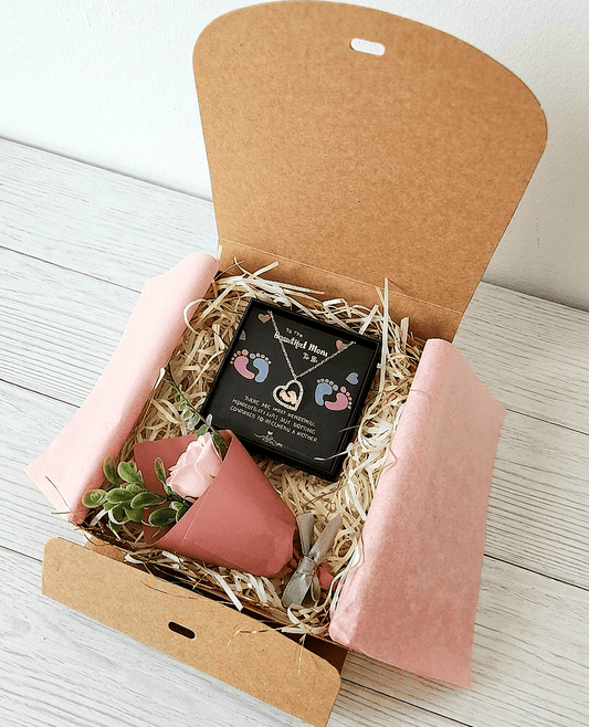 Caja de regalo Kraft abierta vista desde arriba, contiene un colgante en forma de corazón con pies de bebé en su interior sobre una cajita negra con un texto en inglés decorado con huellitas de bebé en rosa y azul, una rosa con envoltorio en rosa palo y lazo gris  sobre una superficie de madera .