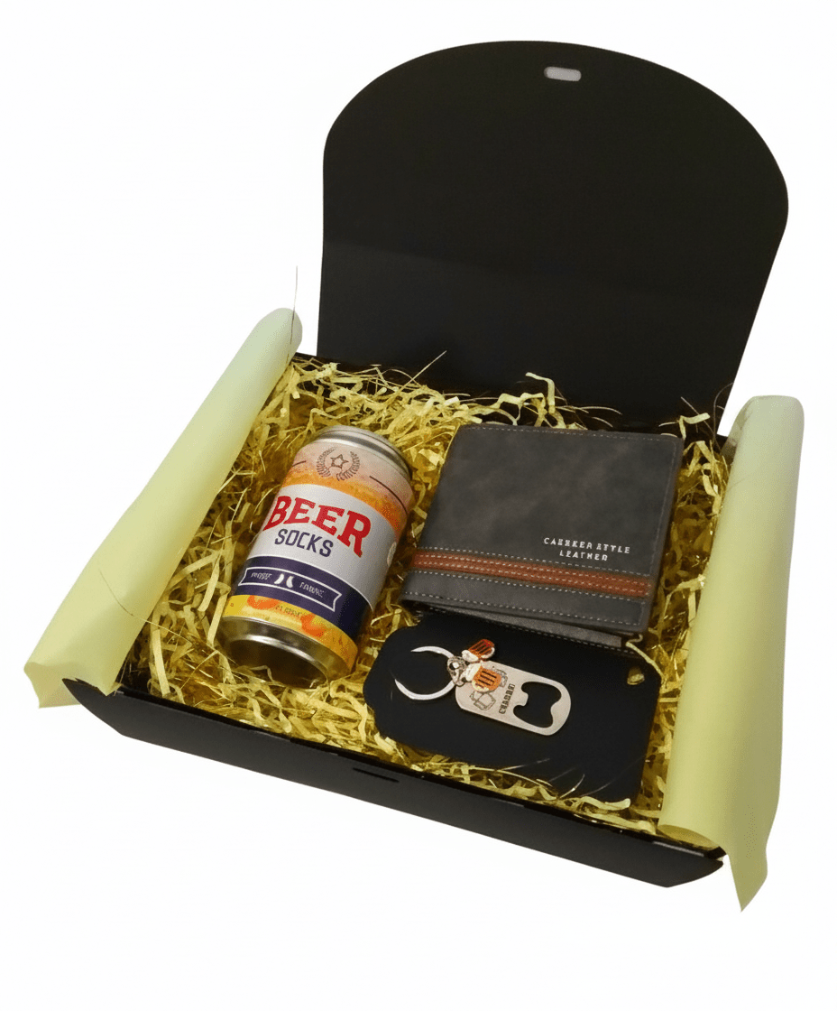 Caja abierta con decoracion de virutas de papel amarillo, mostrando lata de cerveza con calcetines, cartera negra con raya horizontal marrón y llavero de metal.