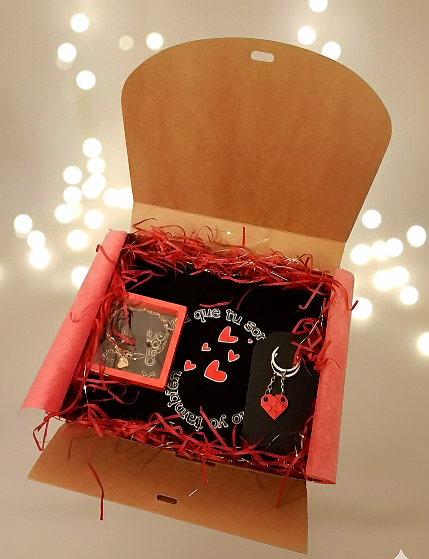 Caja de regalo abierta con virutas de papel rojo que contiene un neceser negro, un  llavero de corazon y un set de joyeria en caja transparente.