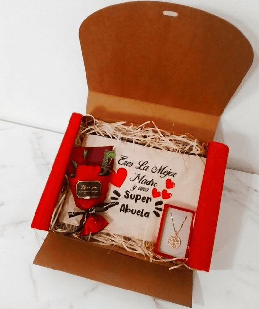 Caja de regalo abierta vista desde arriba, contiene un neceser con frase en español para el dia de la madre, un colgante con el Arbol de la vida en Acero dorado y una rosa con envoltorio rojo y lazo negro, sobre superficie de marmol blanco.