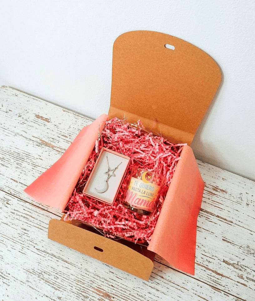 Caja de regalo Kraft vista deste arriba con virutas de papel rosa que contiene una vela con el mensaje  "Te quiero hasta la luna Mamá" y un colgante compuesto por una estrella y media luna todo sobre una superficie de madera blanca.