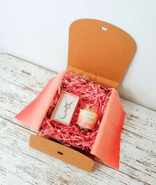 Caja de regalo Kraft vista deste arriba con virutas de papel rosa que contiene una vela con el mensaje  "Te quiero hasta la luna Mamá" y un colgante compuesto por una estrella y media luna todo sobre una superficie de madera blanca.