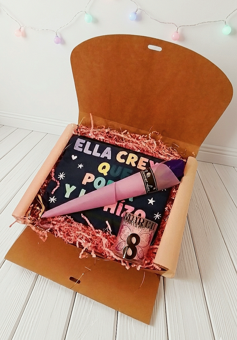 Caja de regalo abierta con virutas de papel rosa, mostrando un neceser con letras multicolor, una vela rosa con diseño drl 8 de Marzo y una rosa morada con envoltorio de color malva sobre superficie de madera blanca y luces led de fondo.