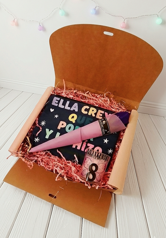Caja de regalo abierta con virutas de papel rosa, mostrando un neceser con letras multicolor, una vela rosa con diseño drl 8 de Marzo y una rosa morada con envoltorio de color malva sobre superficie de madera blanca y luces led de fondo.