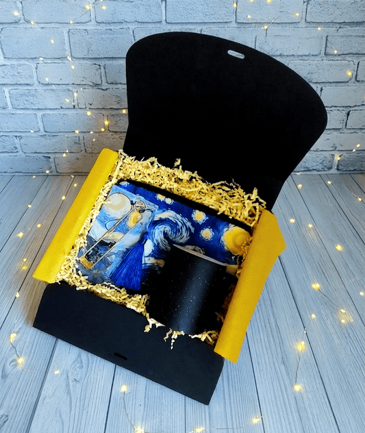 Caja de regalo abierta vista desde arriba donde se muestra en conjunto de regalis compuesto por un neceser con diseño del cuado "La noche estrellada" de Vicent Van Gogh, sobre el neceser hay una taza termosensible de cerámica negra brillante,  a la izquierda puede verse un marcapaginas con adorno azul cobalto presentado en tarjeta con diseño del cuadro "La noche estrellada". La caja se encuentra sobre una superficie de madera azulada y luces led. 