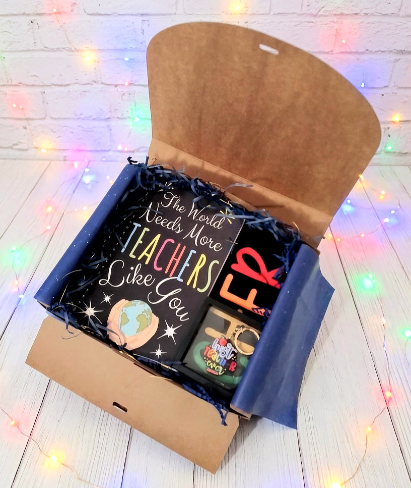 Caja de regalo abierta vista desde arriba, mostrando una agenda negra con espiral de metal y un texto e ilustración con  dos tipos de tipografía y diferentes colores, a su lado puede verse una Bolsa Tote negra plegada donde pueden verse letras de colores, sobre la bolsa hay un llavero de madera y metal con la frase "Best Teacher Ever" con letras de colores presentado en caja de exhibición con marco negro, todo sobre una superficie blanca con luces led de colores.