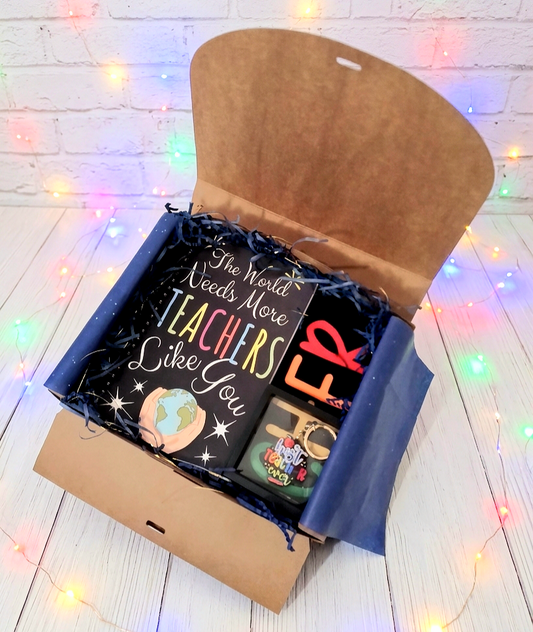Caja de regalo abierta vista desde arriba, mostrando una agenda negra con espiral de metal y un texto e ilustración con  dos tipos de tipografía y diferentes colores, a su lado puede verse una Bolsa Tote negra plegada donde pueden verse letras de colores, sobre la bolsa hay un llavero de madera y metal con la frase "Best Teacher Ever" con letras de colores presentado en caja de exhibición con marco negro, todo sobre una superficie blanca con luces led de colores.