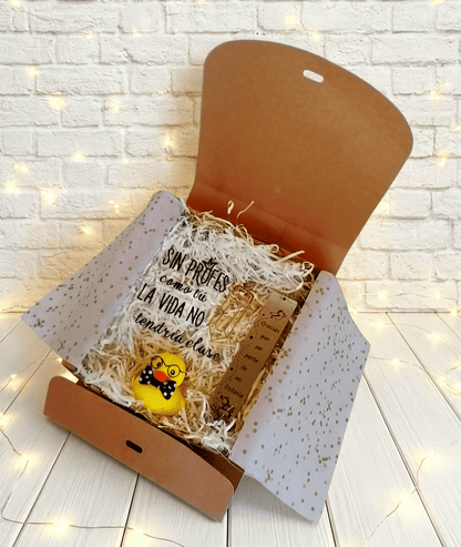 Caja abierta vista desde arriba con un conjunto de artículos de regalo para Profes, compuesto por una Placa Acrilica con la frase "Sin Profes como tu la vida no tendría clase" a su lado en vertical un marcapáginas metálico con un colgate de dije de libro abierto y una frase de agradecimiento para Profesores, por ultimo en la parte inferior cental hay un Patito de Goma amarillo con gafas y pajarita de lunares. La caja se encuentra sobre una superficie de madera blanca con luces led.