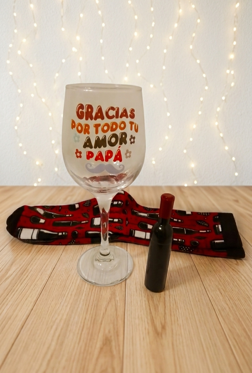 Primer plano de copa de vino personalizada con la frase "GRACIAS POR TODO TU AMOR PAPÁ" junto a sacacorchos con forma de botella de vino y calcetines color burdeos con diseño de botellas y copas sobre fondo de madera clara .