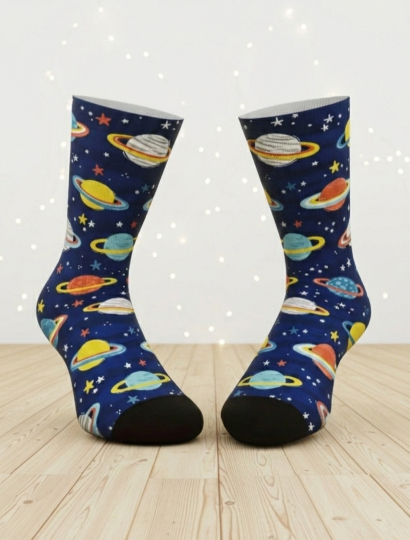 Par de calcetines de color azul marino con un diseño colorido de planetas, anillos de Saturno y estrellas sbre superficie de madera clara y pared blanca con puntos de luz