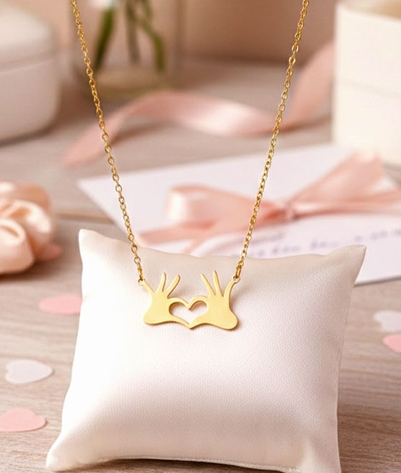 Primer plano de un colgante dorado en Acero Inoxidable de dos manos formando un corazón apoyado sobre un cojin blanco en una superficie de madera.