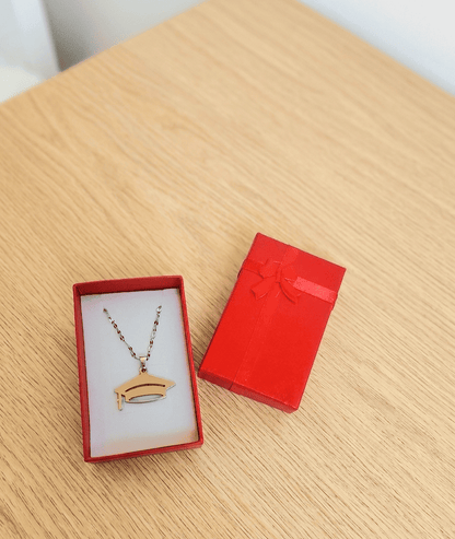 Primer plano de un colgante plateado de Acero inoxidable con un dije de birrete y cadena fina, presentado en una caja de joyeria roja con interior de color blanco,  a su lado se encuentra la tapa de la caja de color rojo decorada con un lazo de raso rojo, todo se encuentra sobre una superficie de madera clara. 