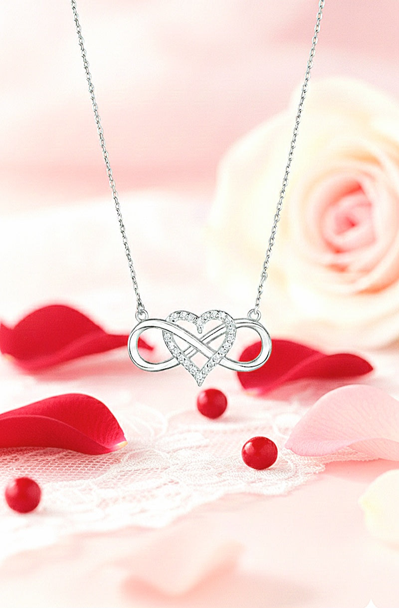 Colgante de Acero con detalle de corazón y simbolo de infinito entrelazados con cristales brillantes presentado en fondo romantico con petalos de rosa rojos y rosas.