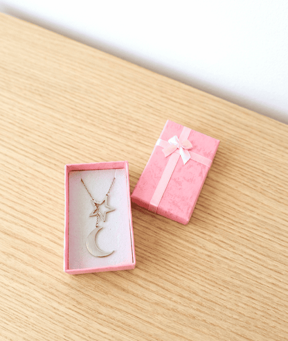 Colgante plateado de Acero inoxidable compuesto por una estrella y media luna entrelazados, colocado en el interior de una cajita de joyeria rosa sobre una superficie de madera clara. 
