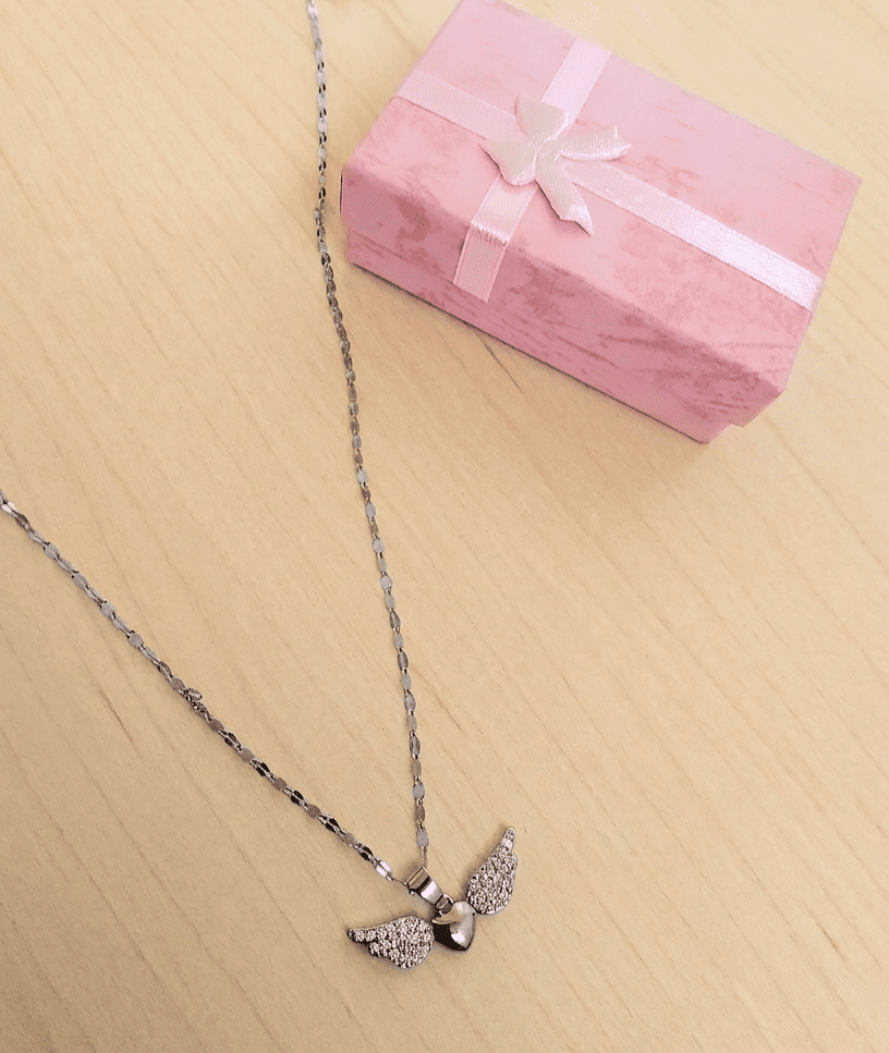 Primer plano visto desde arriba de un colgante de Acero Inoxidable con dije de corazon con alas brillantes y cadena plateada, a su lado puede verse parte de una una cajita de joyería de color rosa, todo sobre una superficie de madera clara. 