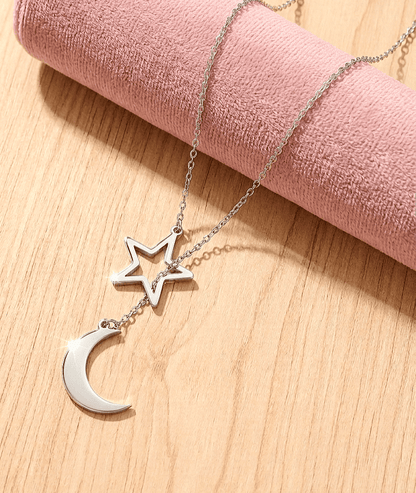 Colgante de cadena fina con una estrella y media luna entrelazados reposando sobre un objeto cilindrico de terciopelo rosa en una superficie de madera clara. 