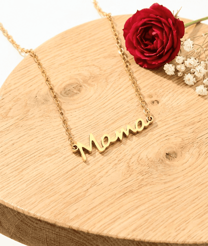 Primer plano de un colgante dorado con la palabra Mamá, destacando sobre una mesa de madera clara junto a una rosa roja con pequeñas flores blancas