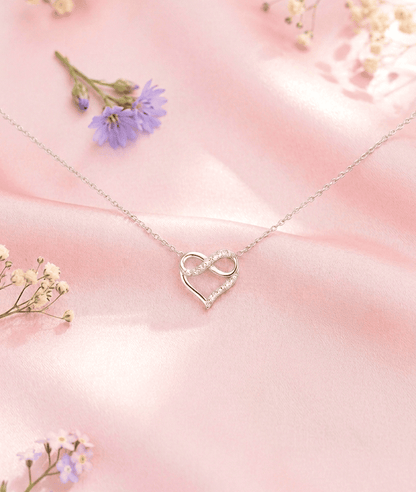 Colgante visto desde arriba compuesto  por un corazón entrelazando el simbolo de infinito adornado con cristales brillantes, sobre un fondo de raso color rosa pastel con unas pequeñas flores silvestres. 