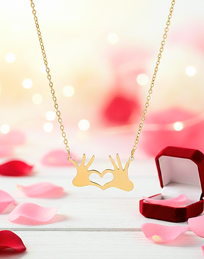 Colgante de Acero dorado con manos formando un corazón con fondo romántico de San Valentín con petalos de rosa.