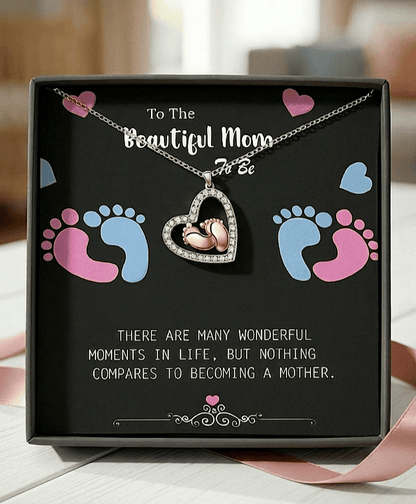 Colgante en forma de corazón con pies de bebé en su interior sobre una caja negra con un texto de dia de la madre en inglés con letras blancas y decorada con huellitas de bebé en rosa y azul.
