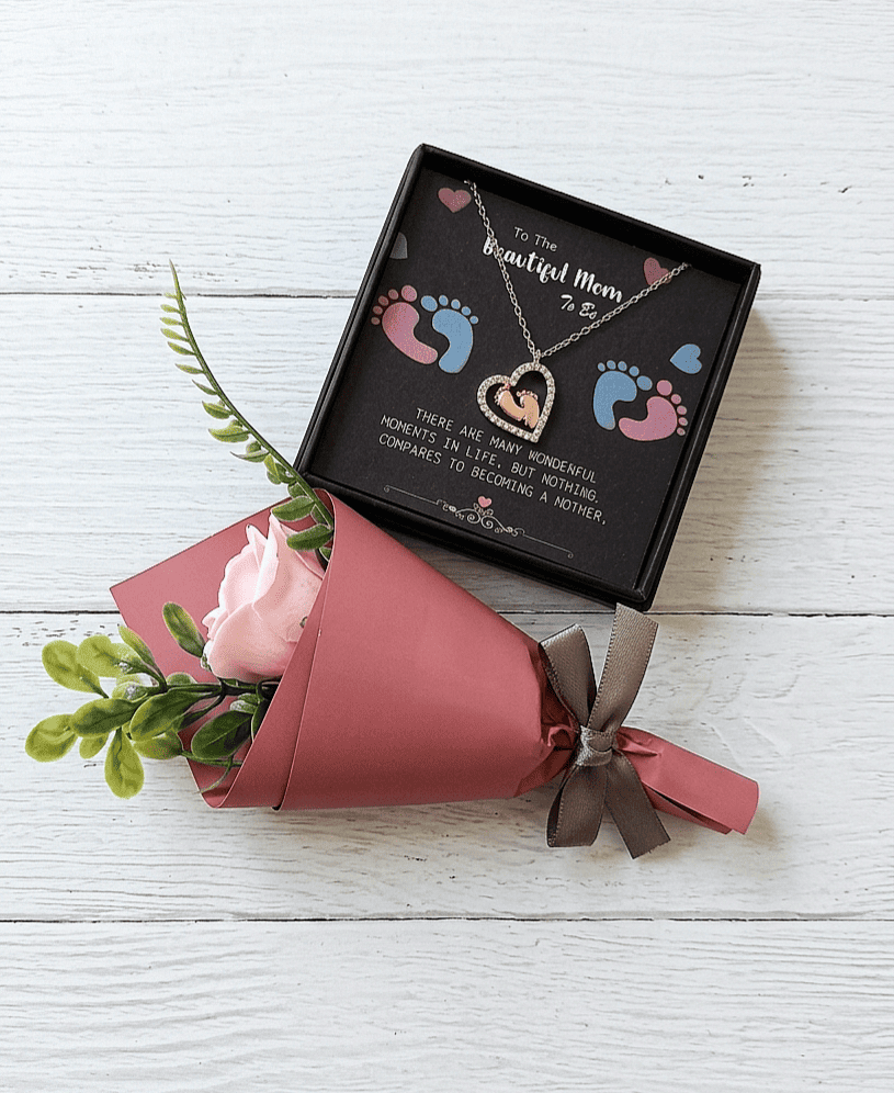 Colgante en forma de corazón con pies de bebe en su interior sobre una caja negra con texto en inglés de dia de la madre con letras blancas, decorada con huellitas de bebé en rosa y azul junto a una rosa con envoltorio de papel y lazo gris sobre superficie de madera blanca. 