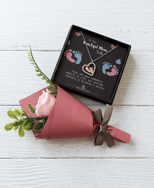 Colgante en forma de corazón con pies de bebe en su interior sobre una caja negra con texto en inglés de dia de la madre con letras blancas, decorada con huellitas de bebé en rosa y azul junto a una rosa con envoltorio de papel y lazo gris sobre superficie de madera blanca. 