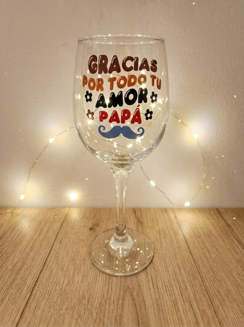 Primer plano de una copa de vino de cristal con la frase " GRACIAS POR TODO TU AMOR PAPÁ " sobre fondo de madera clara y luces decorativas.