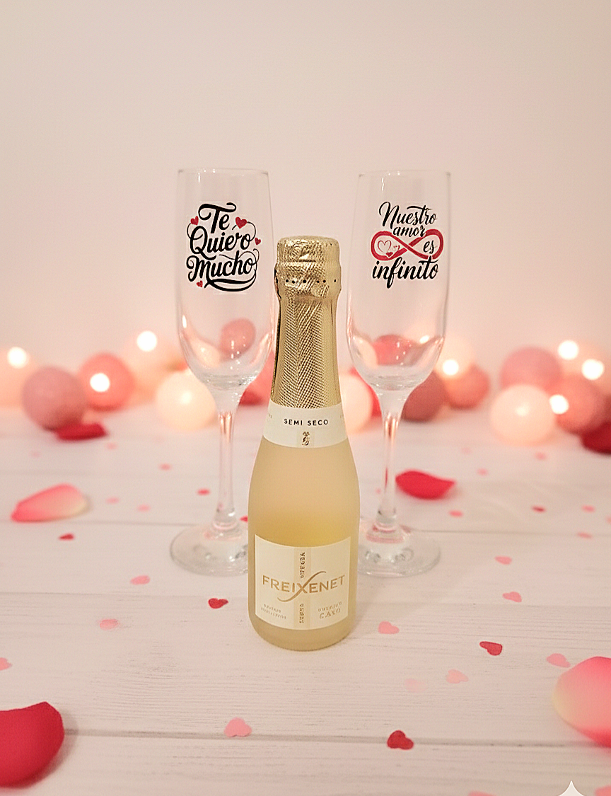Copas de cristal personalizadas con frases en español junto a botella de cava sobre fondo romantico de corazones y petalos de rosa.